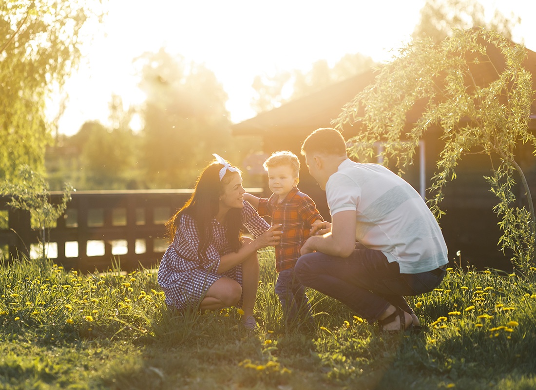 About Our Agency - Portrait of Cheerful Parents Having Fun Spending Time with Their Son Outside on the Green Grass on a Warm Summer Day at Sunset