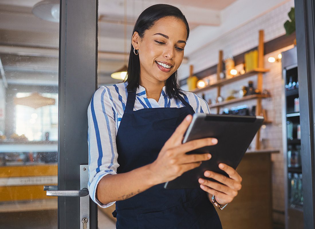 Business Insurance - Closeup Portrait of a Smiling Young Small Business Owner Holding a Tablet While Standing at the Front Door Entrance of her Cafe